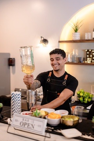 Rey laughing as he pours into a shaker behind the Bar Is Open sign
